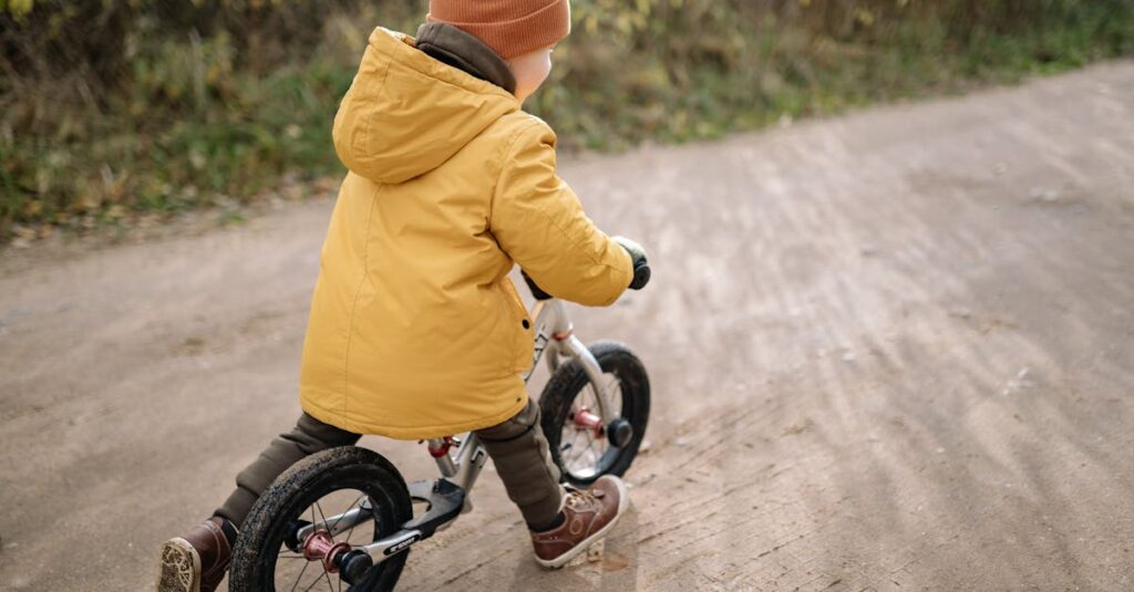 A child wearing a yellow jacket and beanie rides a bike on a dirt path outdoors.