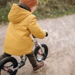 A child wearing a yellow jacket and beanie rides a bike on a dirt path outdoors.