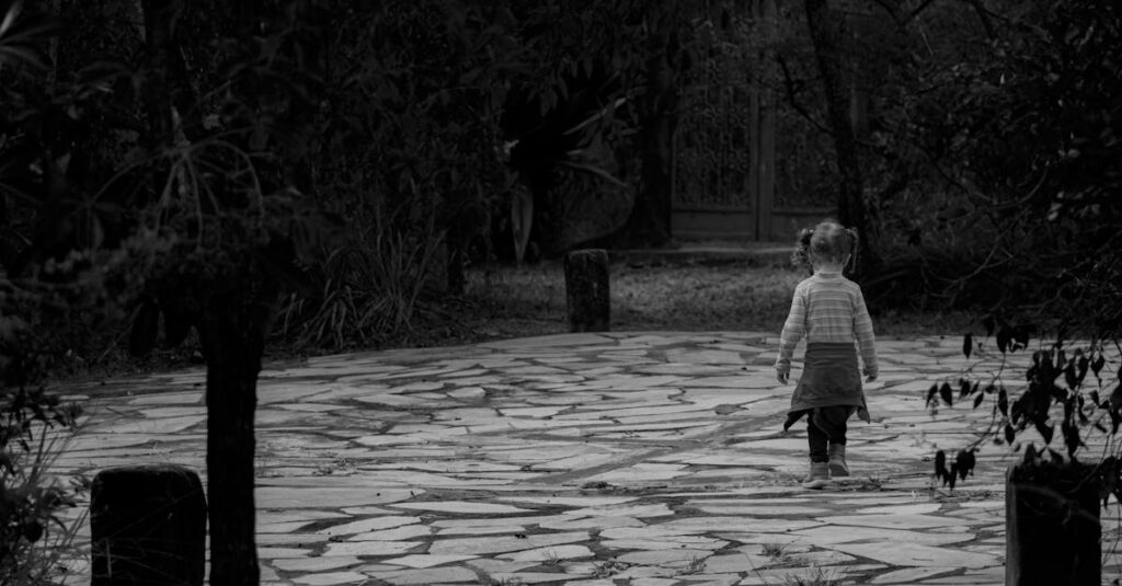 A child with a ponytail walks alone on a stone path surrounded by trees, captured in monochrome.