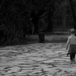 A child with a ponytail walks alone on a stone path surrounded by trees, captured in monochrome.