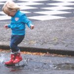 A young child joyfully splashes in a puddle wearing red boots on a rainy day.