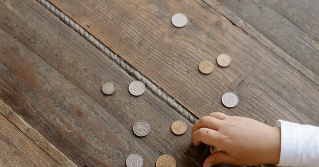 Overhead view of child's hand gathering coins into a glass jar on wooden flooring.