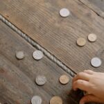 Overhead view of child's hand gathering coins into a glass jar on wooden flooring.