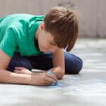 Child in a green shirt drawing on pavement with chalk during the day.