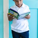 Charming scene of a young boy immersed in reading a book, standing by turquoise doors indoors.