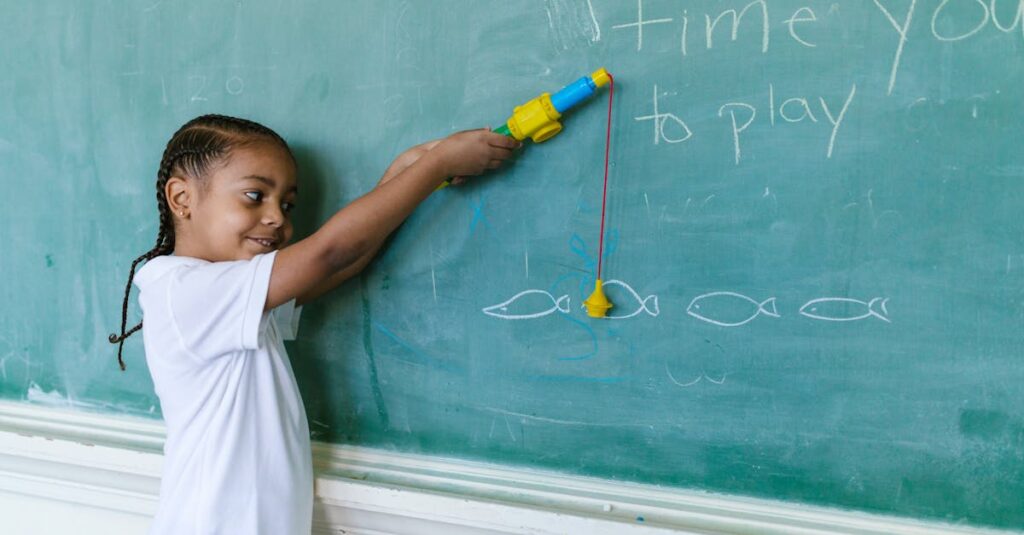 A young boy with braided hair playing with a toy fishing rod by a blackboard in a classroom setting.