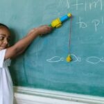 A young boy with braided hair playing with a toy fishing rod by a blackboard in a classroom setting.