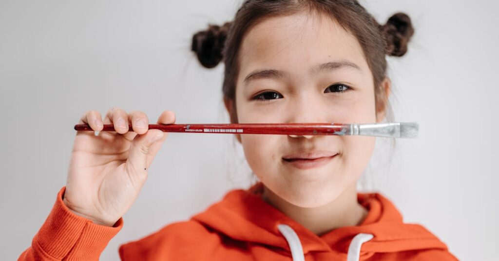 A playful young girl holds a paintbrush under her nose, smiling in a cozy red hoodie.