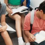 A group of students sitting on stairs, reading and writing with backpacks nearby.