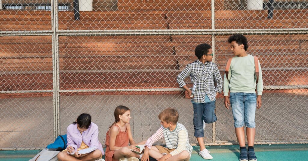 Diverse group of kids sitting and standing by a fence, chatting and studying outdoors.