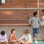 Diverse group of kids sitting and standing by a fence, chatting and studying outdoors.