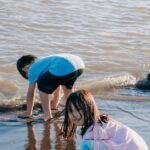 Kids play barefoot on a sunny beach, enjoying the water and sand.
