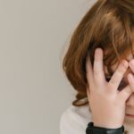 A shy child in a white shirt hiding their face indoors, conveying innocence and introspection.