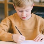 A young boy writing at a table in an art studio with a sign reading 'THERE'S NO PLANET B'.