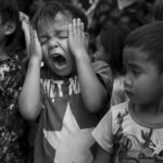 A group of children in a crowd, one appearing distraught, in a black and white setting.