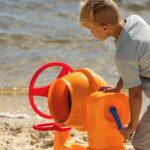 Boy enjoys active play with a toy cement mixer on a sunny lakeside beach in summer.