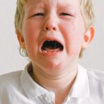 A young child crying, wearing a white shirt, showing strong emotions indoors.