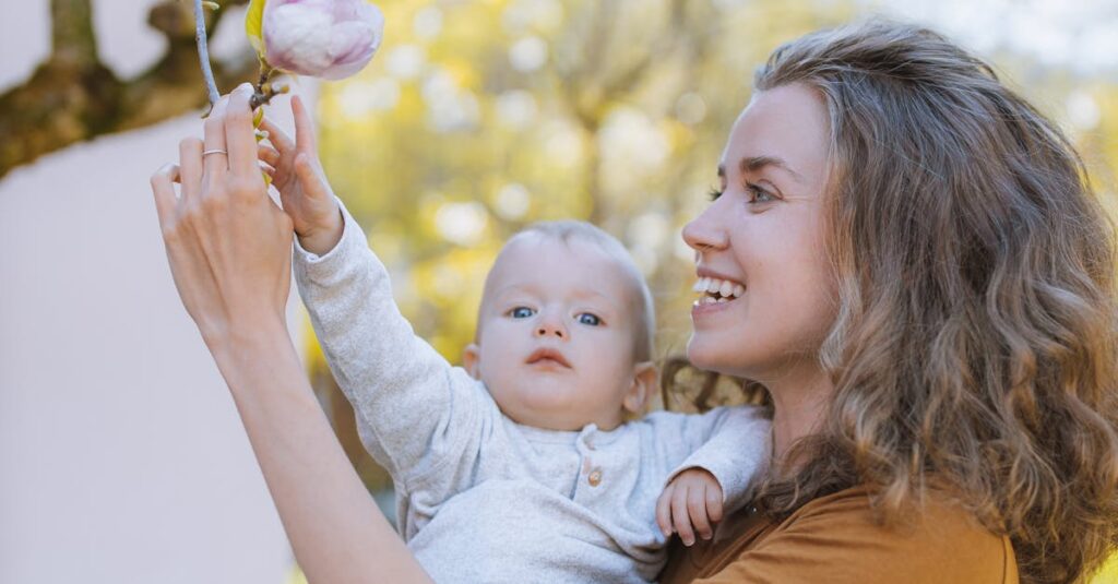 A mother happily holds her baby up to reach blooming magnolias in a sunny garden.