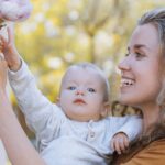 A mother happily holds her baby up to reach blooming magnolias in a sunny garden.