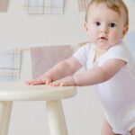 Cute baby standing by a stool indoors with a playful background.