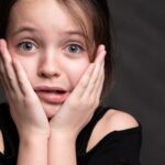 Portrait of a young girl in black top expressing surprise with hands on her face against a dark background.