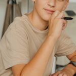 A child with blonde hair pens thoughts in a notebook, appearing thoughtful indoors.