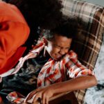 Child in distress on checkered chair with adult leaning over ominously inside a dimly lit room.
