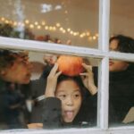 Playful children indoors celebrate Halloween with a pumpkin and festive lights.