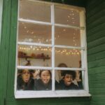 Three joyful children making faces at the window of a cozy cabin, surrounded by autumn scenery.