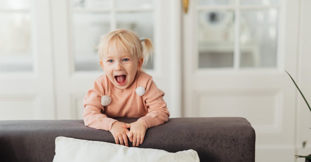 A joyful young girl with blonde hair laughing behind a couch indoors.