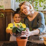 A woman and child enjoy planting a flower together indoors, surrounded by green plants.