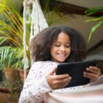 A joyful child relaxing indoors on a hammock while using a tablet, surrounded by plants.