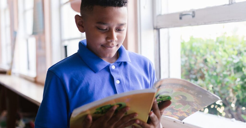 A boy in a blue polo reads a book by a sunlit window, enjoying a quiet moment indoors.