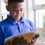 A boy in a blue polo reads a book by a sunlit window, enjoying a quiet moment indoors.