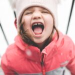 Cheerful child wearing winter clothing screams happily in the snowy outdoors.