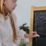 Concentrated schoolgirl standing near easel blackboard and writing mathematical equation in cozy light room