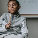 Concentrated African American boy in casual outfit sitting on chair with pencil and copybook and thinking how to do task
