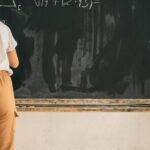 Back view full body of clever faceless girl writing on blackboard while solving mathematical equation during lesson in light room